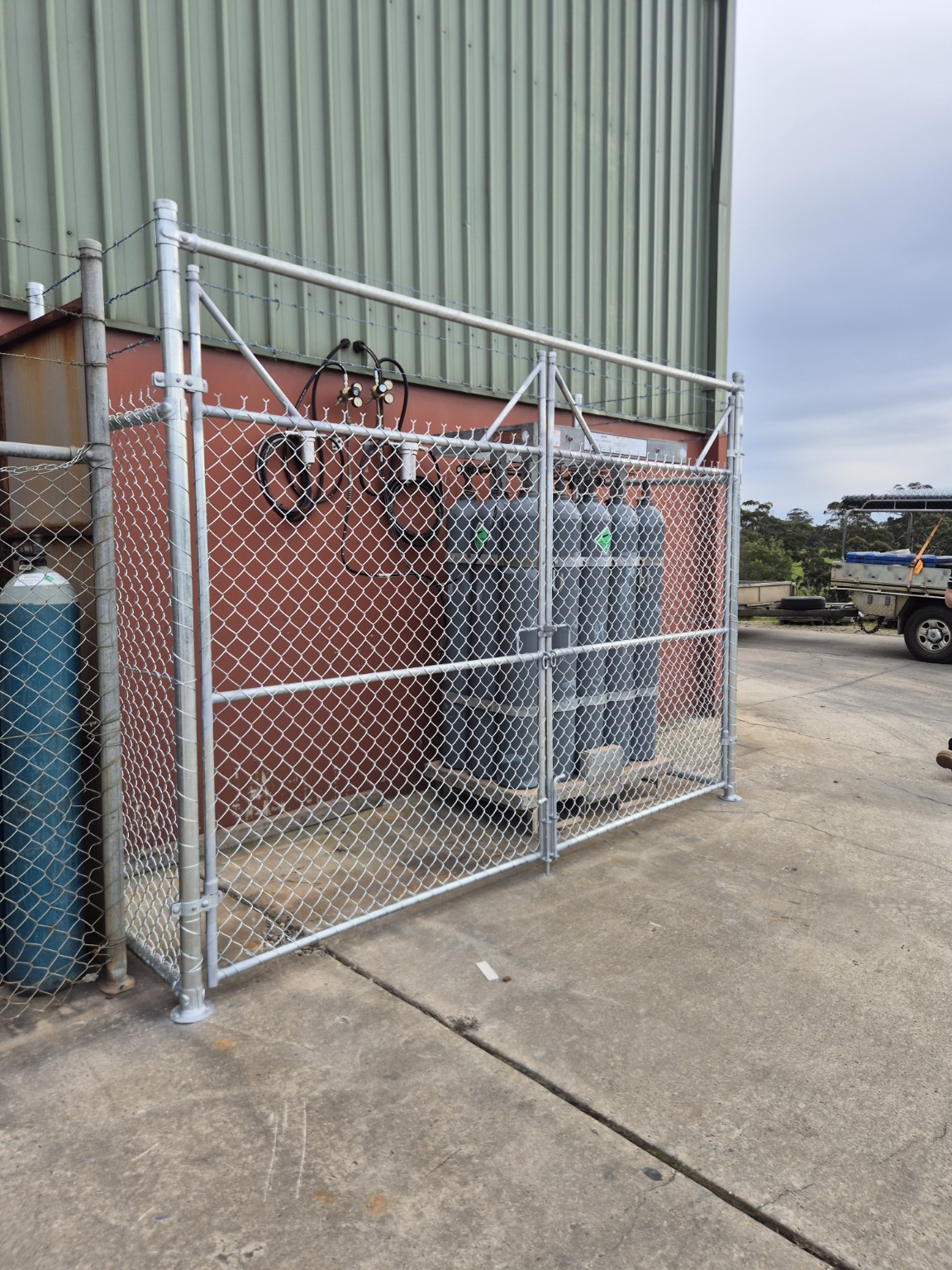 Close view of paired galvanised chain mesh gates and barbed wire around existing gas cylinders